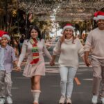 A joyful family walks down a decorated street wearing Christmas hats, symbolizing holiday spirit.