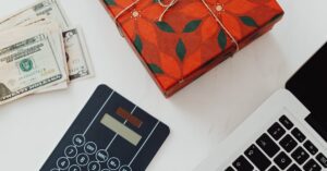 A beautifully wrapped gift box, cash, calculator, and laptop on a white table, symbolizing holiday expenses.