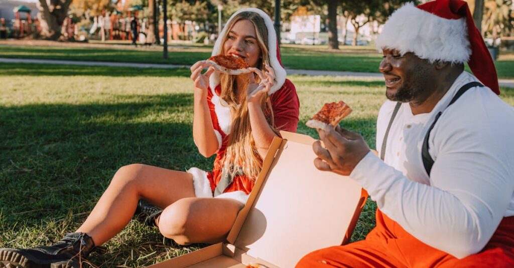 A joyful duo in Santa costumes enjoy pizza in a park, capturing festive fun.