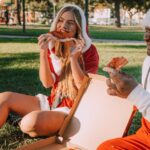 A joyful duo in Santa costumes enjoy pizza in a park, capturing festive fun.