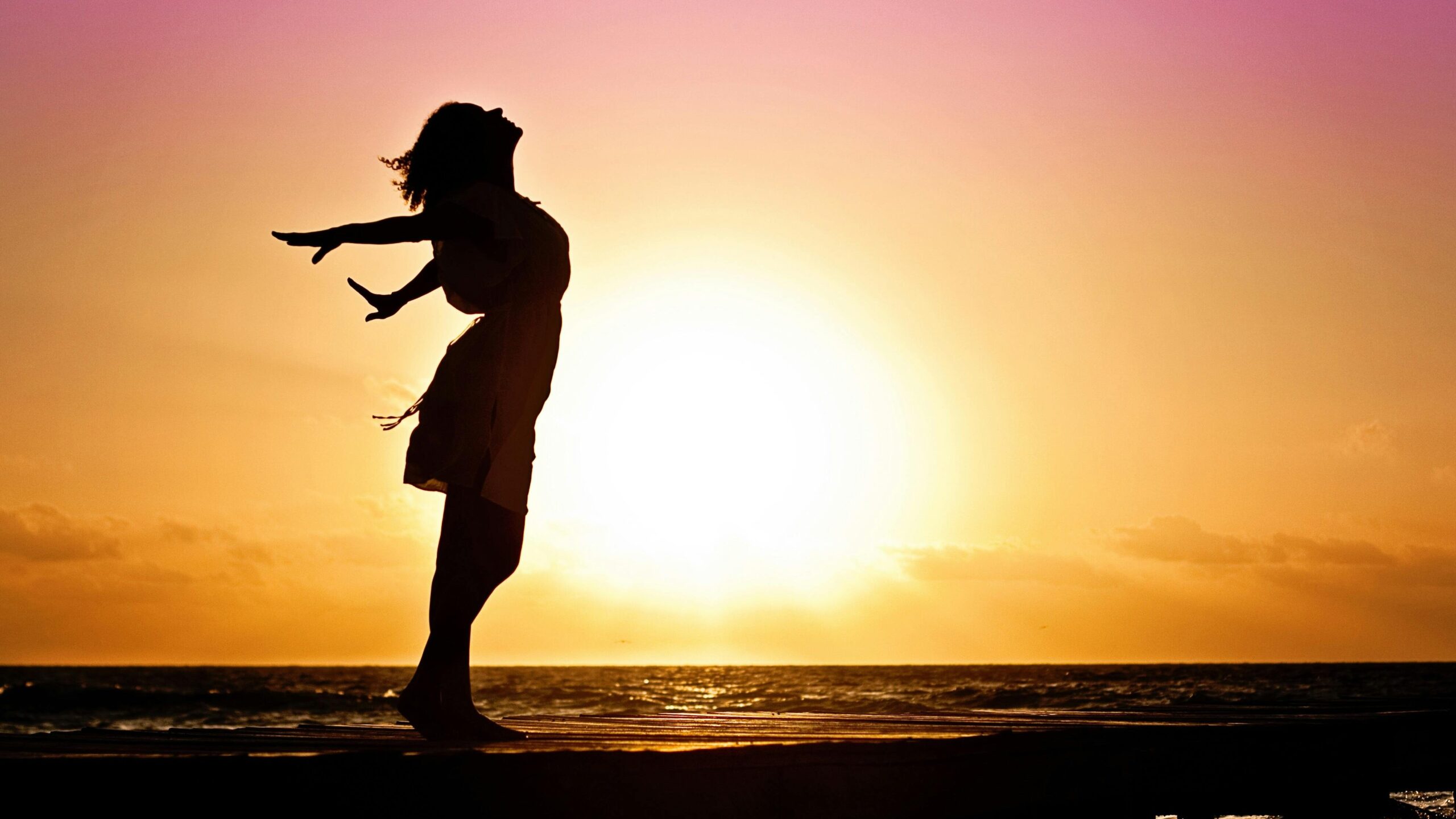 Silhouette of a woman at the beach with arms outstretched against a vibrant sunset backdrop.