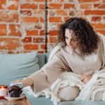 Woman with curly hair enjoys coffee on a cozy sofa, wrapped in a knitted blanket.