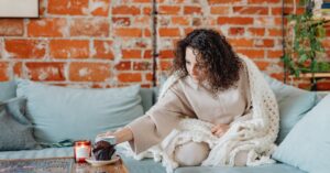 Woman with curly hair enjoys coffee on a cozy sofa, wrapped in a knitted blanket.