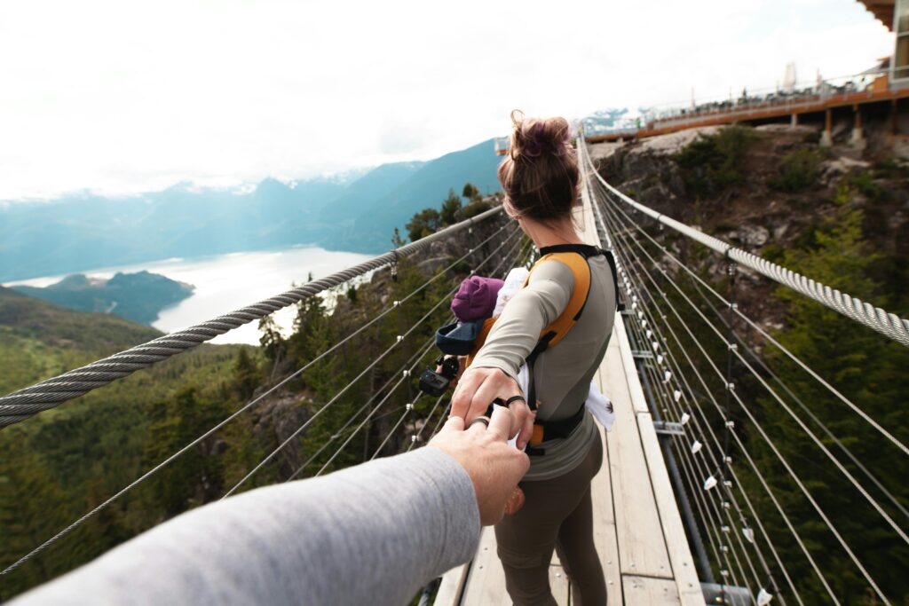 A woman with a baby crosses a scenic hanging bridge, holding hands amid a mountainous landscape.