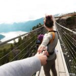 A woman with a baby crosses a scenic hanging bridge, holding hands amid a mountainous landscape.